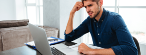 Picture of confused young man sitting near laptop and holding head with his hand. Looking at laptop., Credit: Stock Image