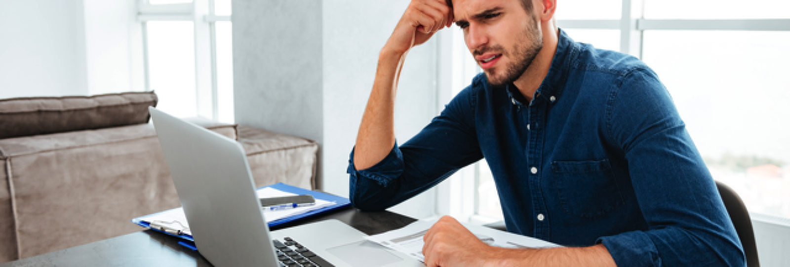 Picture of confused young man sitting near laptop and holding head with his hand. Looking at laptop., Credit: Stock Image